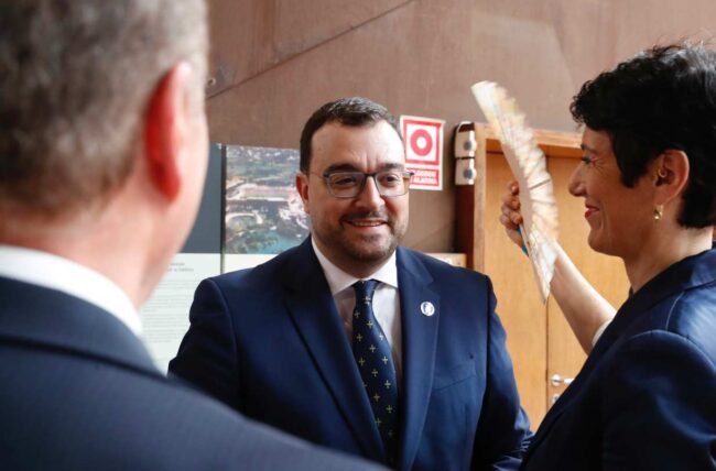 Barbón apuesta por una nueva etapa para Asturias – NB Radio Digital Man in a navy suit and glasses smiles as a woman hands him a document in a hallway, with another person’s head in the foreground on the left.